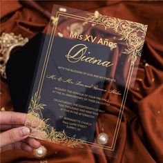 a person holding up a black and gold wedding card in front of a red cloth