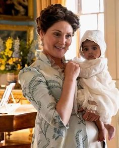 a woman holding a baby wearing a white dress and head scarf in front of a window