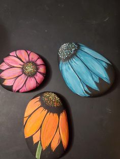 three painted rocks with flowers on them sitting on a black surface next to each other