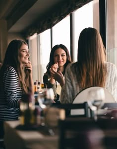 three women sitting at a table talking to each other and drinking wine in front of a window