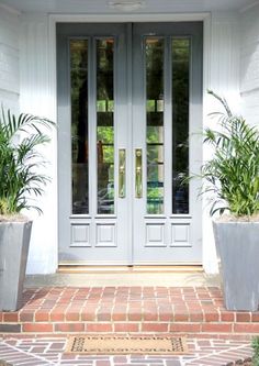 two potted plants sitting on the side of a white building with double doors and brick walkway