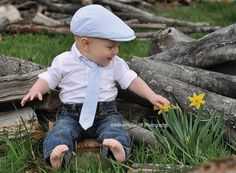 a little boy sitting in the grass wearing a tie and a hat with a daffodil