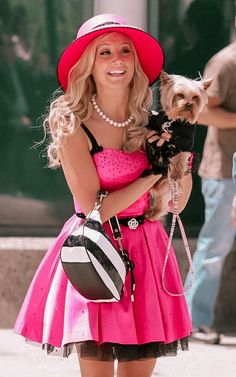 a woman in a pink dress and hat holding a small dog while walking down the street