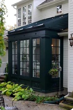 a small house with a black roof and green trim around the windows, sitting on gravel in front of a white house