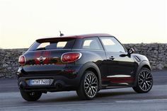 the rear end of a small black car parked in front of a stone wall and fence