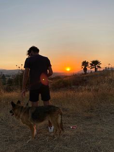 a man standing next to a dog on top of a dry grass field at sunset