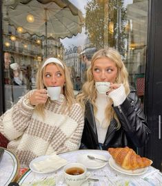 two women sitting at a table with cups of coffee and croissants in front of them