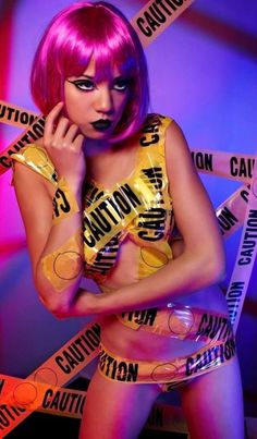 a woman with bright pink hair and black makeup posing in front of caution tape wrapped around her body