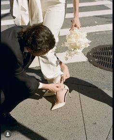 a man kneeling down next to a woman in white shoes and holding a wedding bouquet