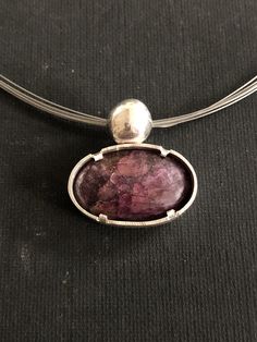 A beautiful purple Sunstone 1 1/4 x 3/4 inches)with a gorgeous sparkly shimmer is set in a solid Sterling Silver pendant. On top of the pendant is a big casted solid Sterling Silver pebble. On back of the pebble is the loop for the necklace - it seems the pendant is floating on the wire necklace! The rim of the thick bezel is left a bit rough to emphasize the character of the stone. A great little pendant with a fun and unique character! T Silver Necklace With Large Oval Pendant, Silver Necklace With Large Oval Cabochon Stone, Purple Sterling Silver Necklace With Large Pendant, Purple Cabochon Round Pendant Jewelry, Purple Pendant Jewelry With Large Stone, Artistic Purple Pendant Jewelry, Amethyst Pendant With Large Stone, Wire Necklace, Sterling Silver Pendants