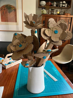 a vase filled with paper flowers sitting on top of a blue cutting mat next to a wooden table