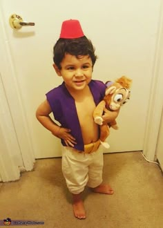 a young boy holding a stuffed animal in front of a door wearing a red hat