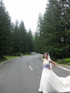 a woman in a wedding dress standing on the side of a road with trees behind her