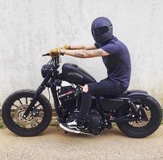 a man riding on the back of a black motorcycle down a street next to a white wall