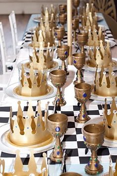 a table topped with lots of gold cups and plates covered in crowns on top of checkered cloth