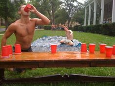 a man sitting at a picnic table with cups in front of him drinking from a red cup