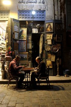 two men sitting at a table in front of a store with pictures on the wall