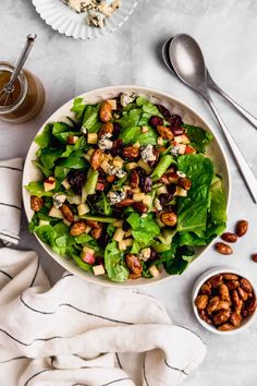 a white bowl filled with salad and nuts next to two silver spoons on a table