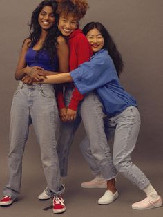 three young women hugging each other in front of a gray background with one woman smiling and the other leaning on her back