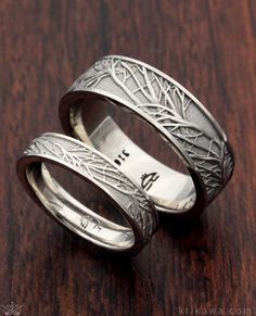 two wedding rings with engraved designs on them sitting on a wooden surface, one is silver and the other is white gold