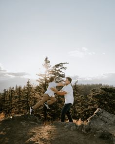 two people jumping up into the air on top of a hill with trees in the background