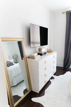 a bedroom with a dresser, mirror and television on it's stand in front of a window