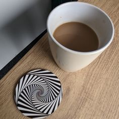 a cup of coffee sitting next to a coaster on a wooden table with a black and white design