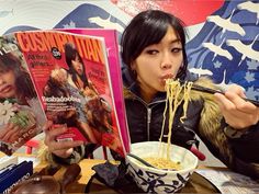 a woman eating noodles from a bowl with chopsticks and reading the magazine's cover