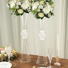 two clear vases filled with white flowers sitting on top of a wooden table next to candles