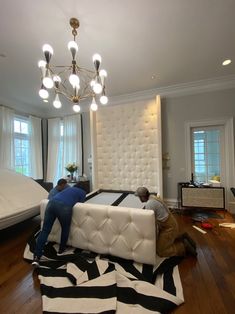 two men working on a bed in a room with hardwood floors and white walls,