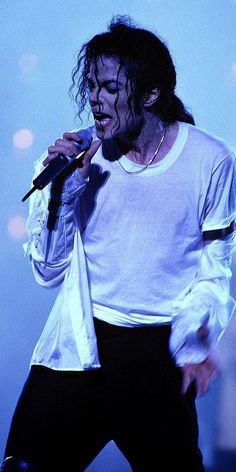 a man in white shirt and black pants on stage holding a microphone to his mouth