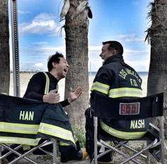 two firemen sitting on folding chairs talking to each other in front of palm trees