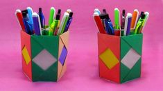 two colorful pens and pencils are in a paper holder on a pink surface,