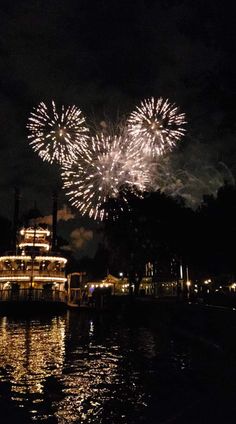 fireworks are lit up in the night sky over a boat on water with buildings and lights behind it