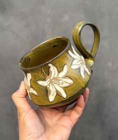 a hand holding a green and white vase with flowers on it's side, in front of a gray background