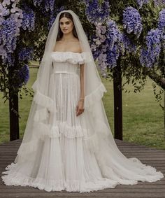 a woman in a white wedding dress standing on a wooden platform with purple flowers behind her