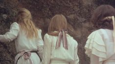 three women in white dresses standing next to each other near a stone wall and holding hands