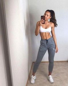 a woman taking a selfie in her white top and grey sweatpants, while looking at her cell phone