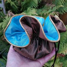 a hand holding a brown and blue purse in front of some green plants on the ground