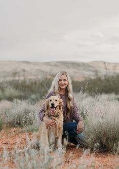 a woman sitting in the middle of a field with her dog and smiling at the camera