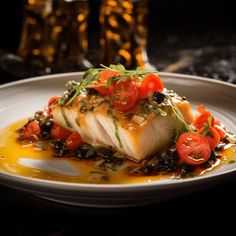 a white plate topped with fish covered in tomatoes and olives next to a glass of beer