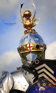 a close up of a knight's helmet on top of a metal object with clouds in the background