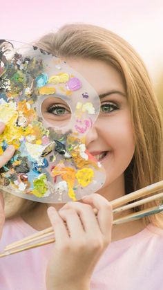 a woman holding up a plate with paint and brushes in front of her face as she holds chopsticks