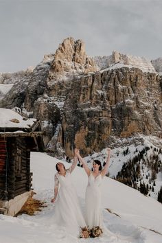 two brides standing in the snow holding their hands up with mountains in the background