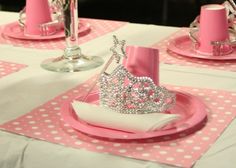 a table set with pink plates, napkins and silver tiara on top of them