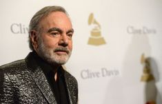 an older man with grey hair and beard wearing a black jacket standing in front of a white wall