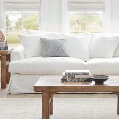 a living room with white couches and pillows on the coffee table in front of two windows