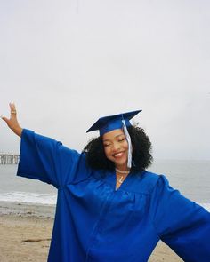 a woman wearing a blue graduation gown and holding her arms out in the air while standing on a beach