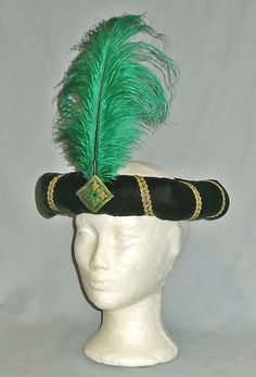 a white mannequin head wearing a black hat with green and gold feathers on it