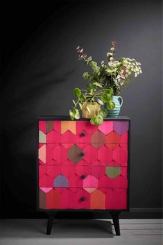 a colorful chest of drawers with flowers and plants on top in front of a black wall